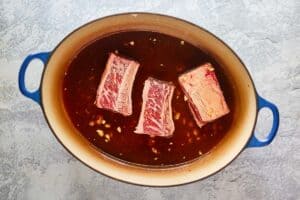 three raw beef short ribs and Asian sauce in a pot.