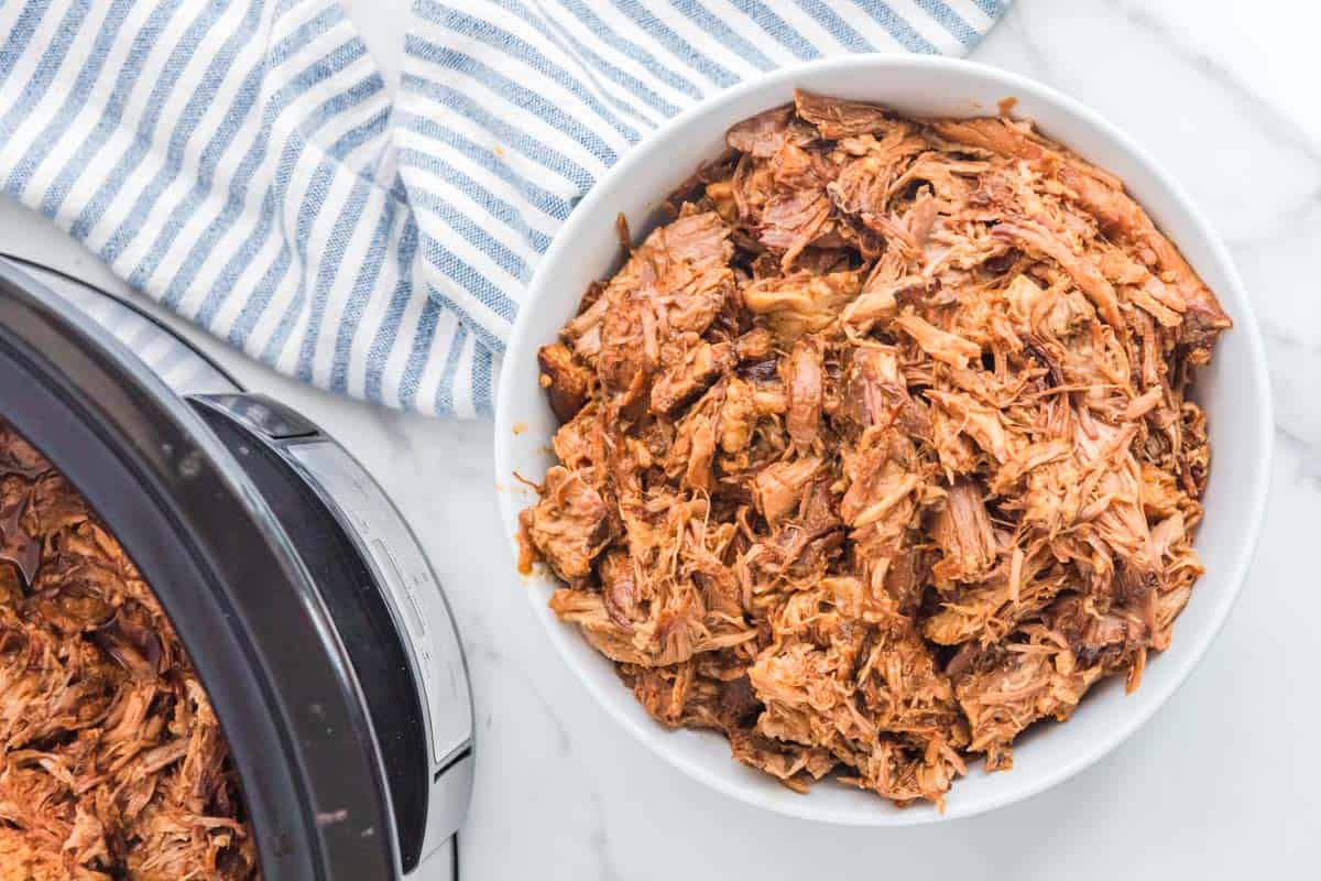 Slow cooker pulled pork in a bowl next to a crockpot with the BBQ pork in it.