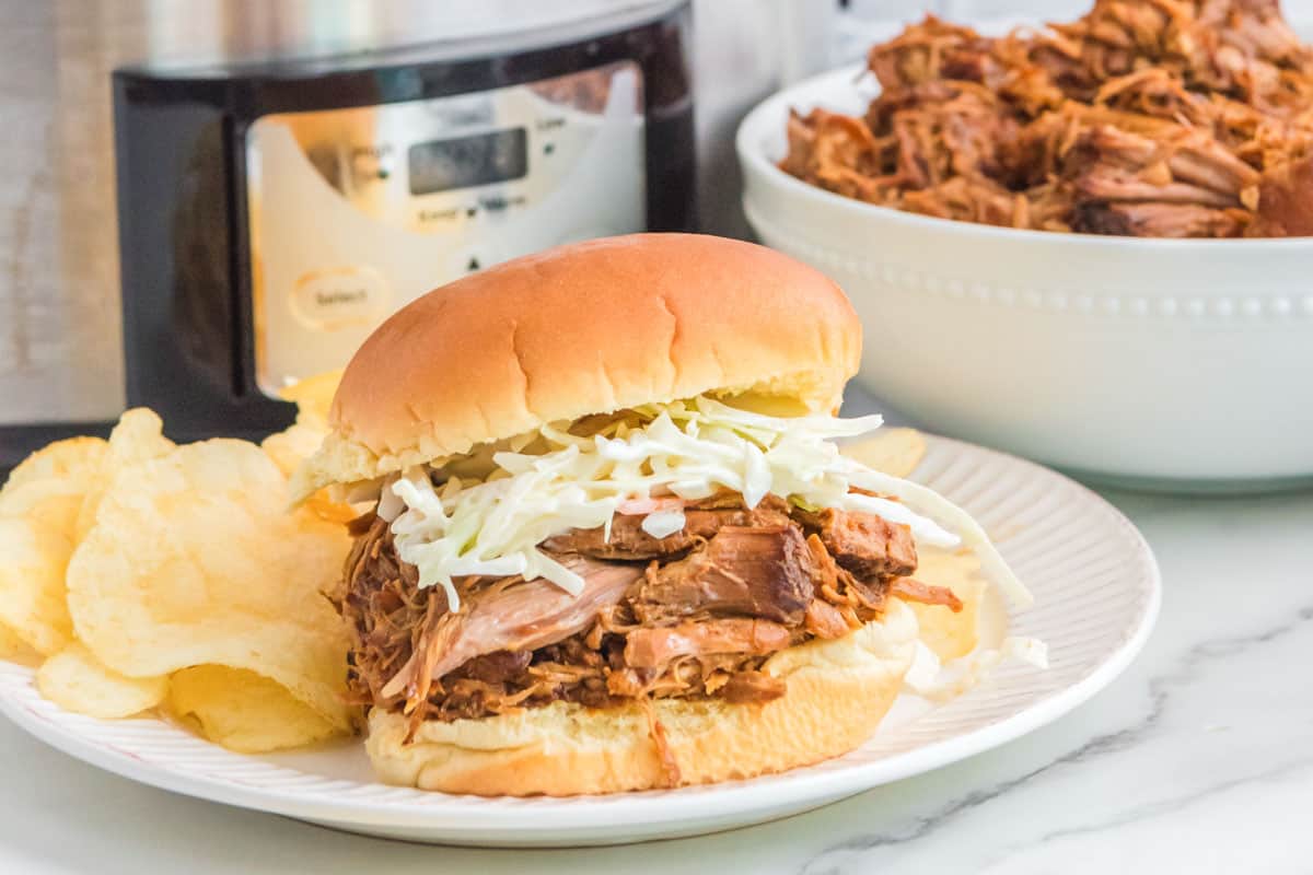 Slow cooker BBQ pulled pork sandwich with coleslaw, potato chips, and a bowl of the shredded pork.