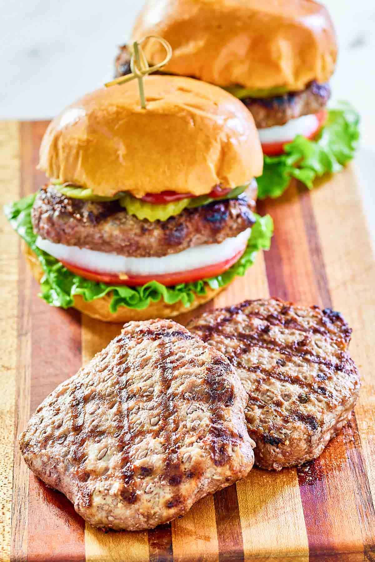 Two grilled frozen burger patties and two burgers on a wood serving board.