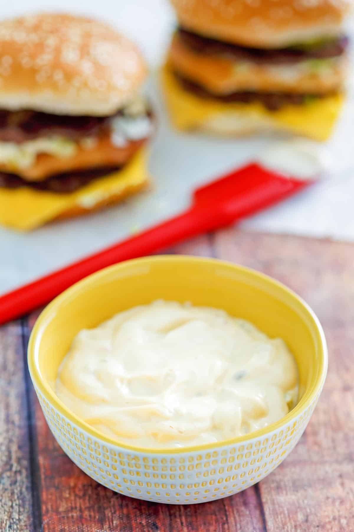 Burger sauce in a bowl and two burgers behind it.